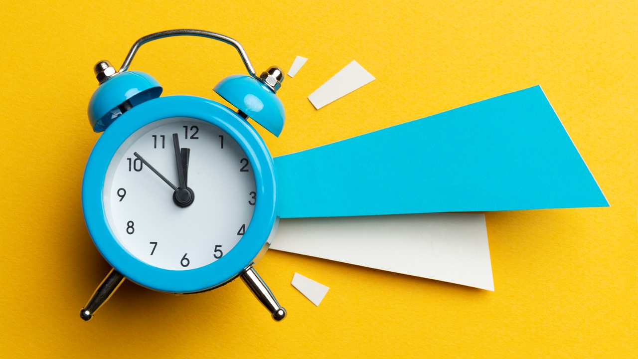 A blue alarm clock on a yellow background. Blue and white shapes are to the left of the clock for emphasis. The clock shows the time of 11:50 and the shapes make it look like the alarm is going off. 